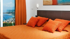 Un dormitorio con una cama grande, ropa de cama naranja, vista al mar y un cuadro abstracto colorido en la pared.