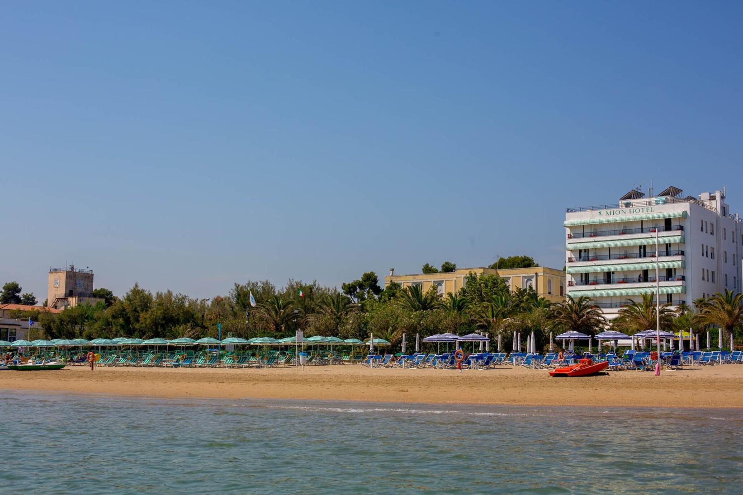 Una playa con hileras de sombrillas y sillas de descanso, un edificio con la leyenda 'MION HOTEL' al fondo, y un cielo azul despejado.
