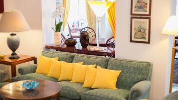 Ein grünes Sofa mit gelben Kissen, ein kleiner Tisch mit einem blauen Gegenstand, eine Lampe und eine Vase mit Blumen.