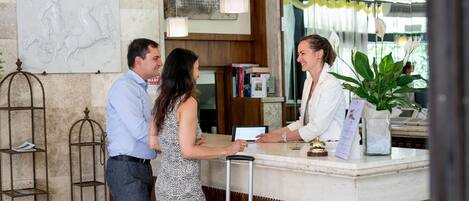 A reception desk with a receptionist, a man, and a woman standing in front of it. There is a plant on the desk, a metal shelf with books, and a map on the wall.