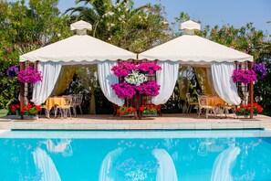 A poolside area with two covered seating areas, each with a table and chairs, surrounded by lush greenery and flowering plants. - LH Mion Hotel (Silvi)