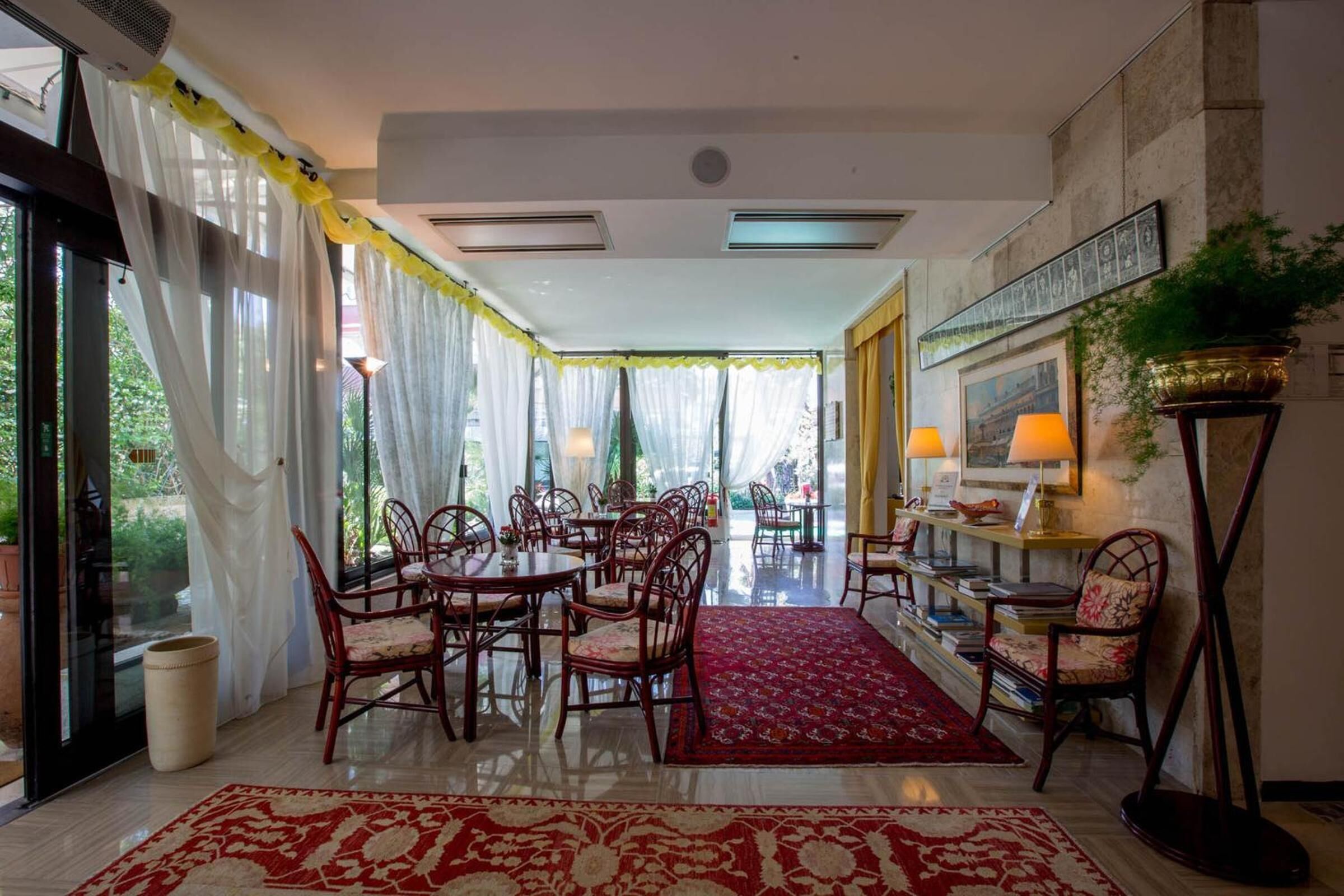 Un comedor amplio con mesas y sillas redondas, una alfombra estampada roja, y vistas al exterior a través de ventanas grandes.