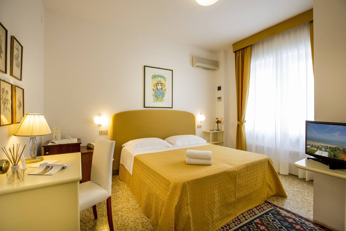 A hotel room with a bed, a desk with a lamp, a television, and framed artwork on the wall.