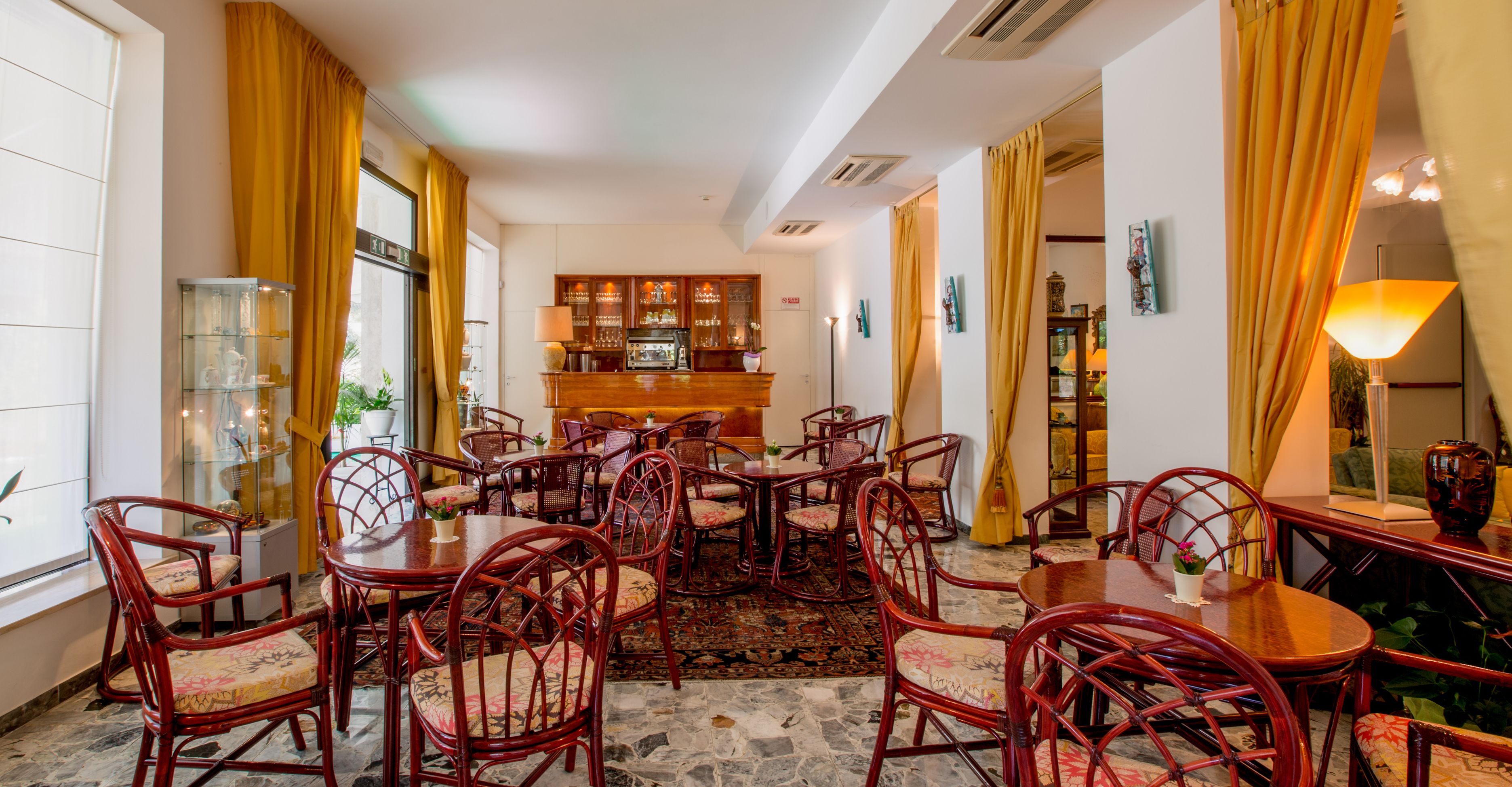 A spacious dining area with multiple wooden tables and chairs, a bar in the background, and large windows with curtains.