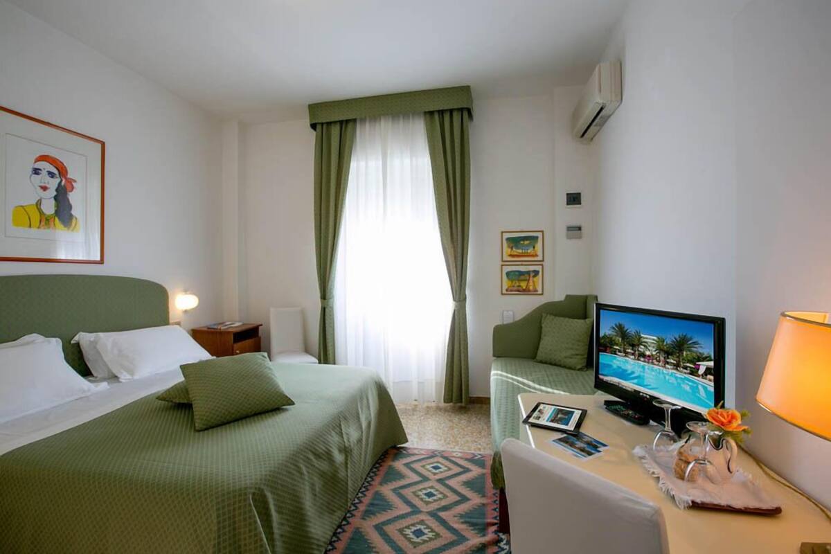 A hotel room with a bed, a desk with a computer, a TV displaying a pool, and a window with curtains.