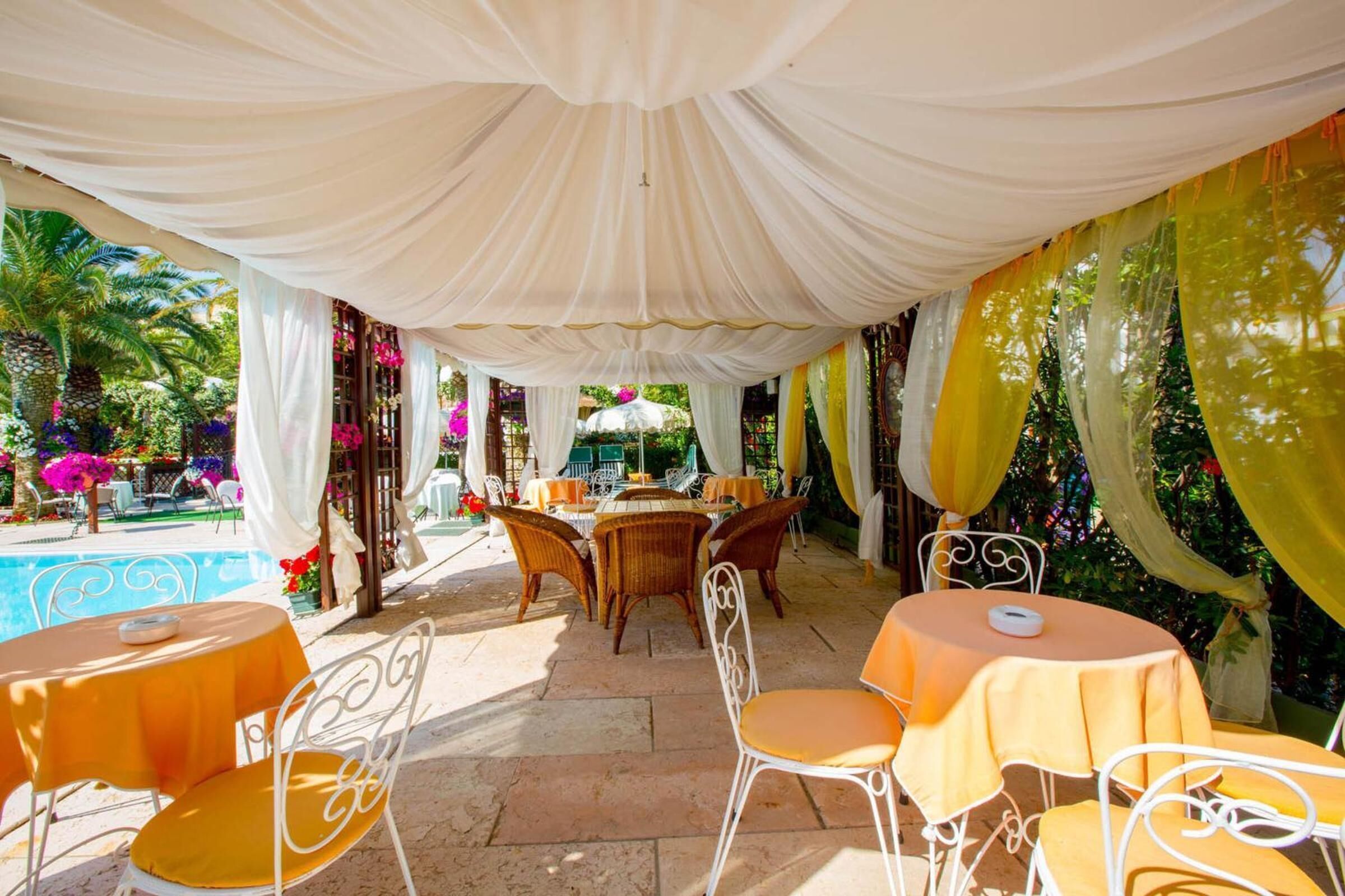 Un área de comedor al aire libre con sillas y mesas de metal blanco, adornada con tela blanca y amarilla.