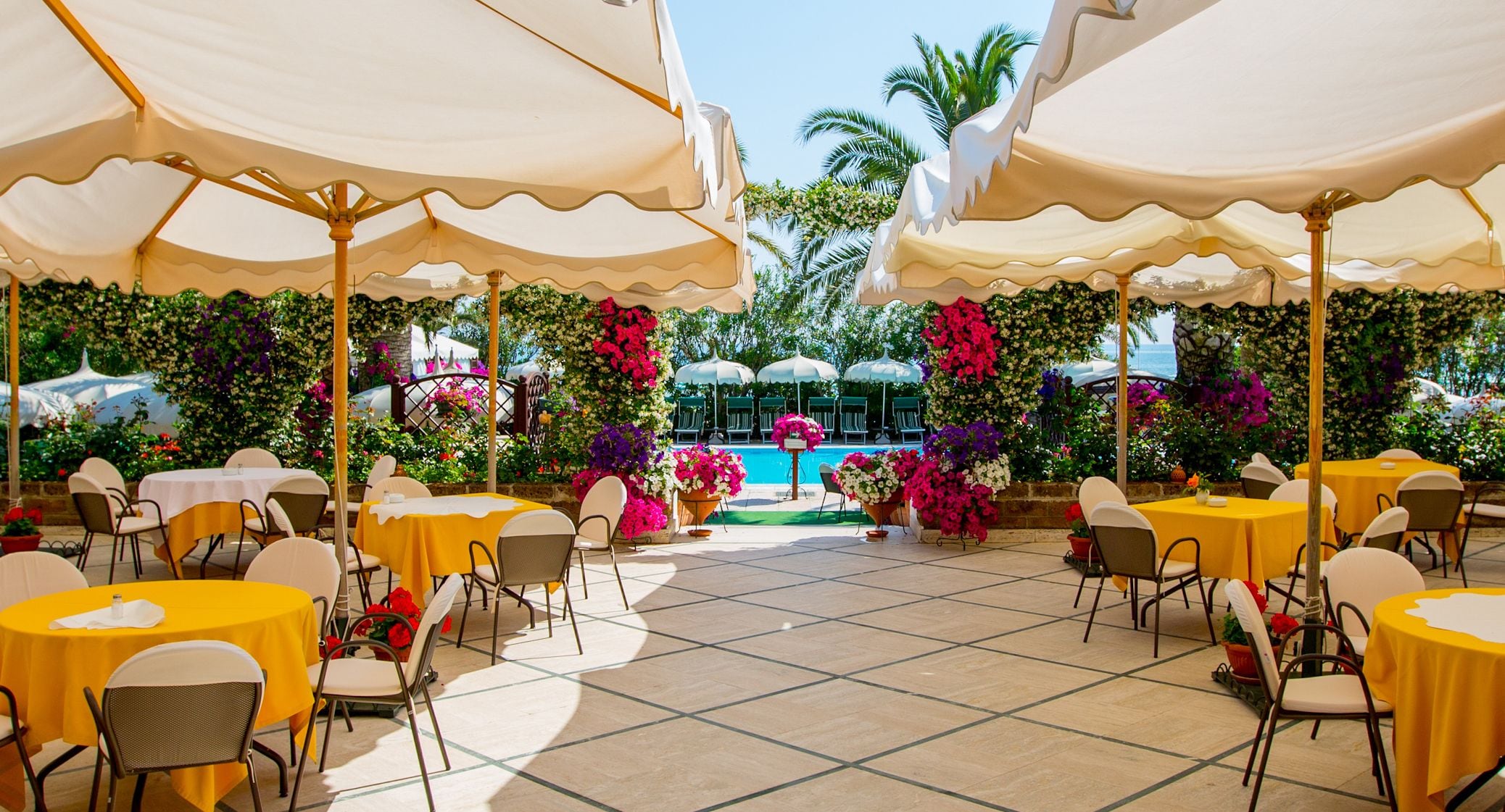 An outdoor dining area with yellow tables and chairs, white umbrellas, and a pool in the background.