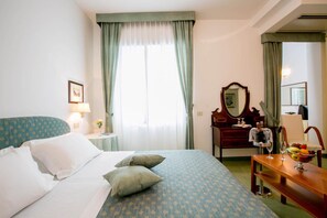 A hotel room with a bed, a wooden dresser, a mirror, a desk with a bowl of fruit, and a window with curtains. - LH Mion Hotel (Silvi)