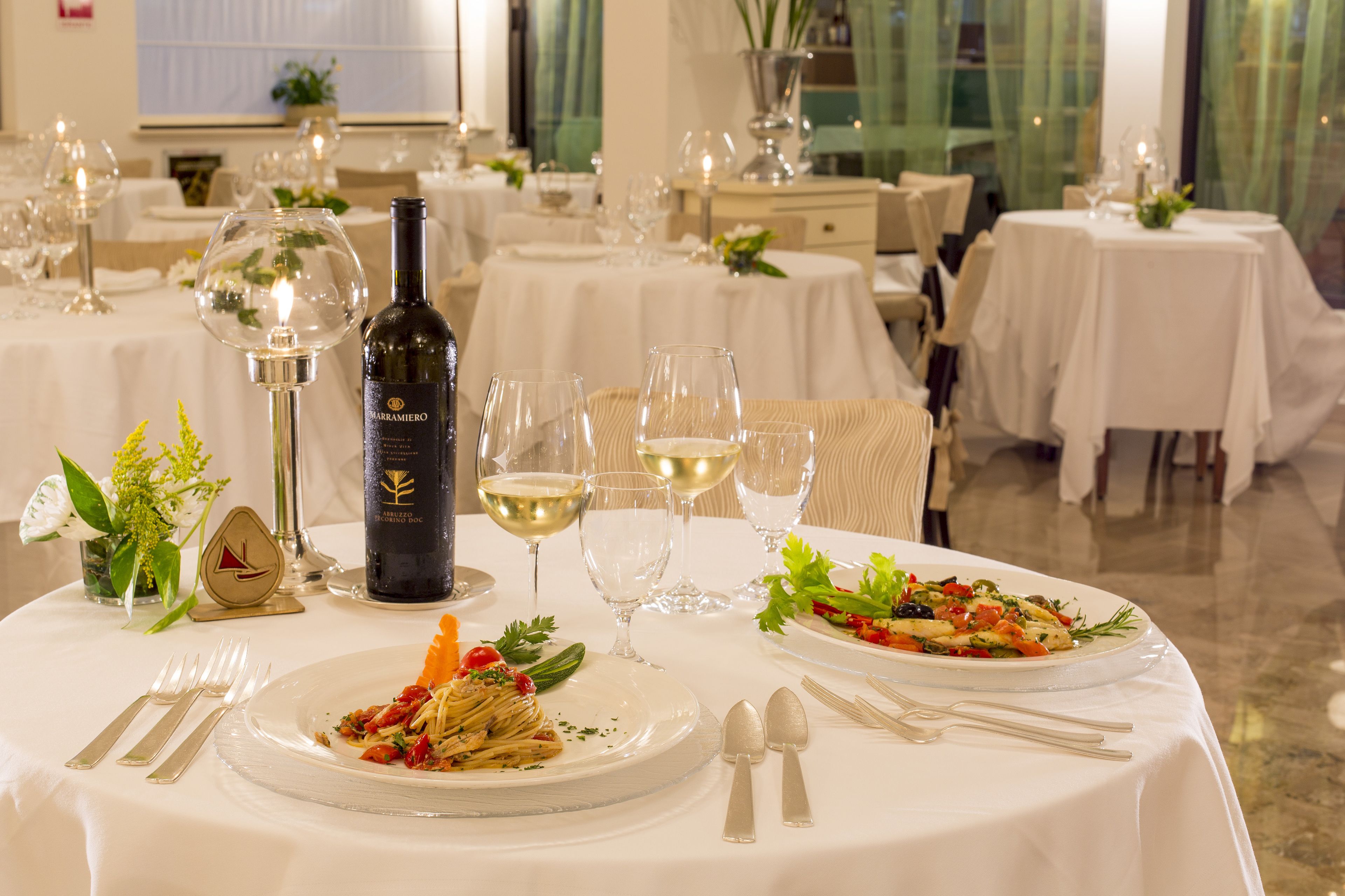 A restaurant setting with multiple tables, each set with white tablecloths, wine glasses, and plates of food.
