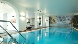 An indoor swimming pool with a waterfall mural, clear glass walls, and a modern design. - Hotel Tanneck (Bad Woerishofen)