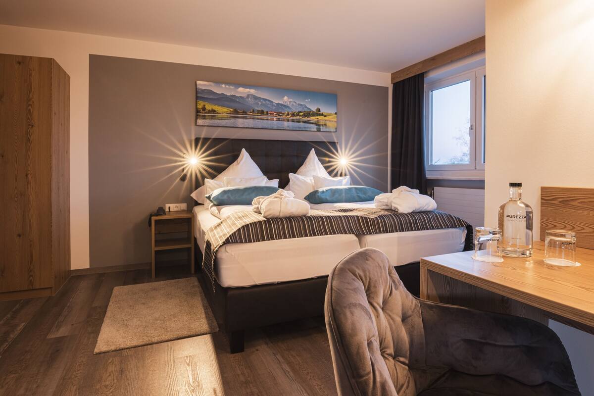 Une chambre d’hôtel avec un lit, un bureau, une chaise, une table de chevet, une fenêtre et un tableau au mur.