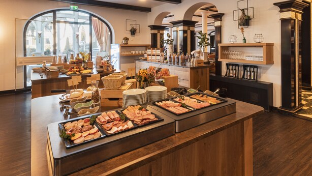 A buffet setup with various food items and beverages, a seating area with chairs, and a large window providing natural light. - Hotel Tanneck (Bad Woerishofen)