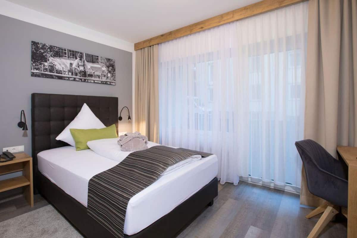 Une chambre d’hôtel avec un grand lit, un bureau, une chaise, une table de chevet et une fenêtre avec des rideaux légers.