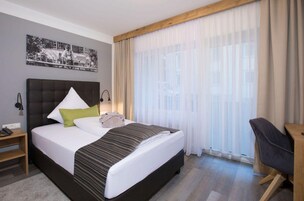 Habitación de hotel con una cama grande, un escritorio, una silla, una mesita de noche y una ventana con cortinas translúcidas.