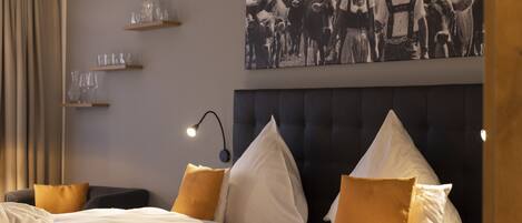 A hotel room with a large bed, multiple pillows, a headboard, a wall-mounted shelf with glasses, and a framed black and white photograph above the bed.