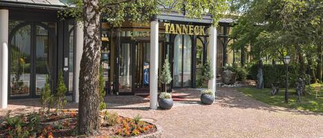 A Tanneck hotel entrance with a brick pathway, potted plants, and a clear sky.