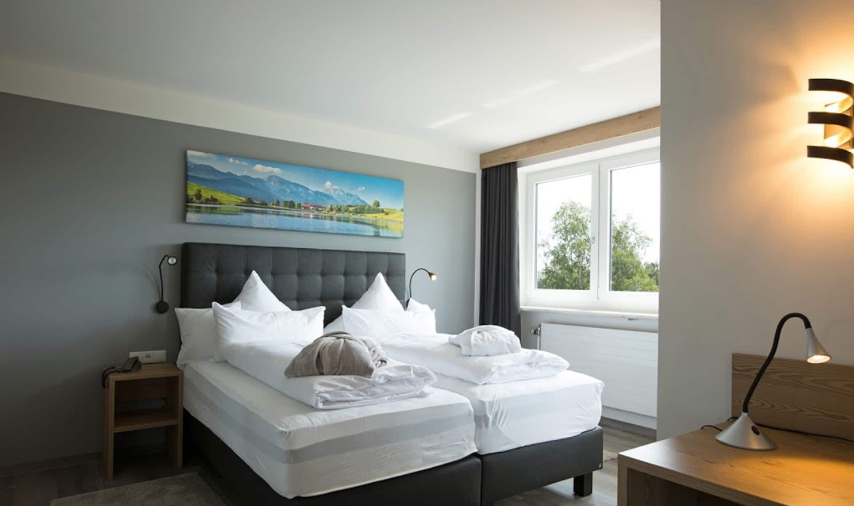A modern hotel room with a large bed, a bedside table, a nightstand, a painting of a landscape, and a window with curtains.