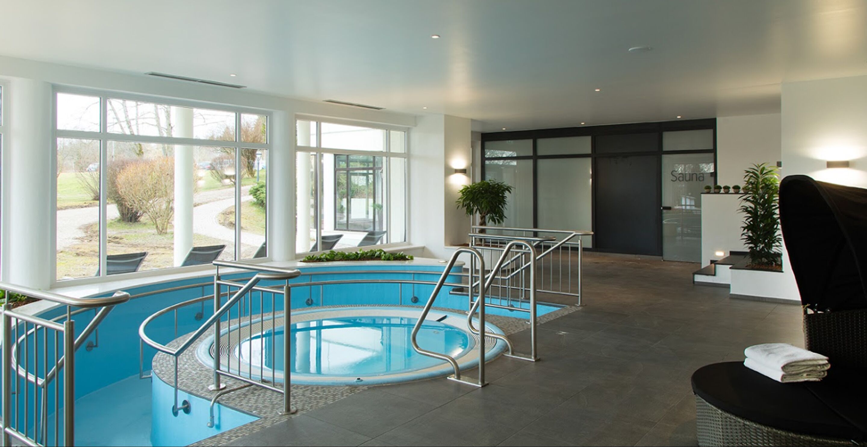A modern indoor swimming pool with clear glass walls, a sunlit area with seating, and a reception desk.
