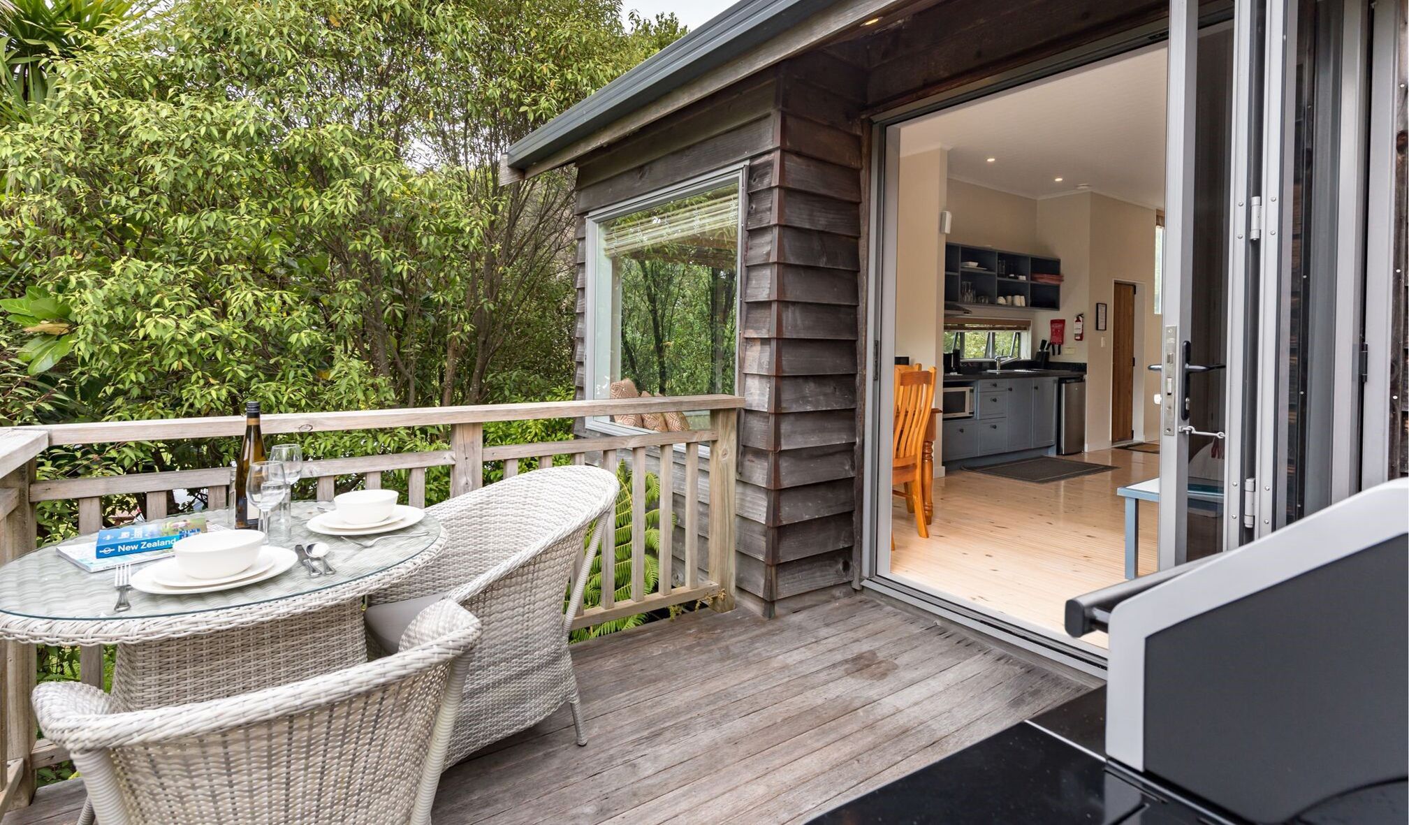 Une terrasse en bois, meublée de meubles en osier, avec une table en verre, et donnant sur une cuisine moderne par une porte ouverte.