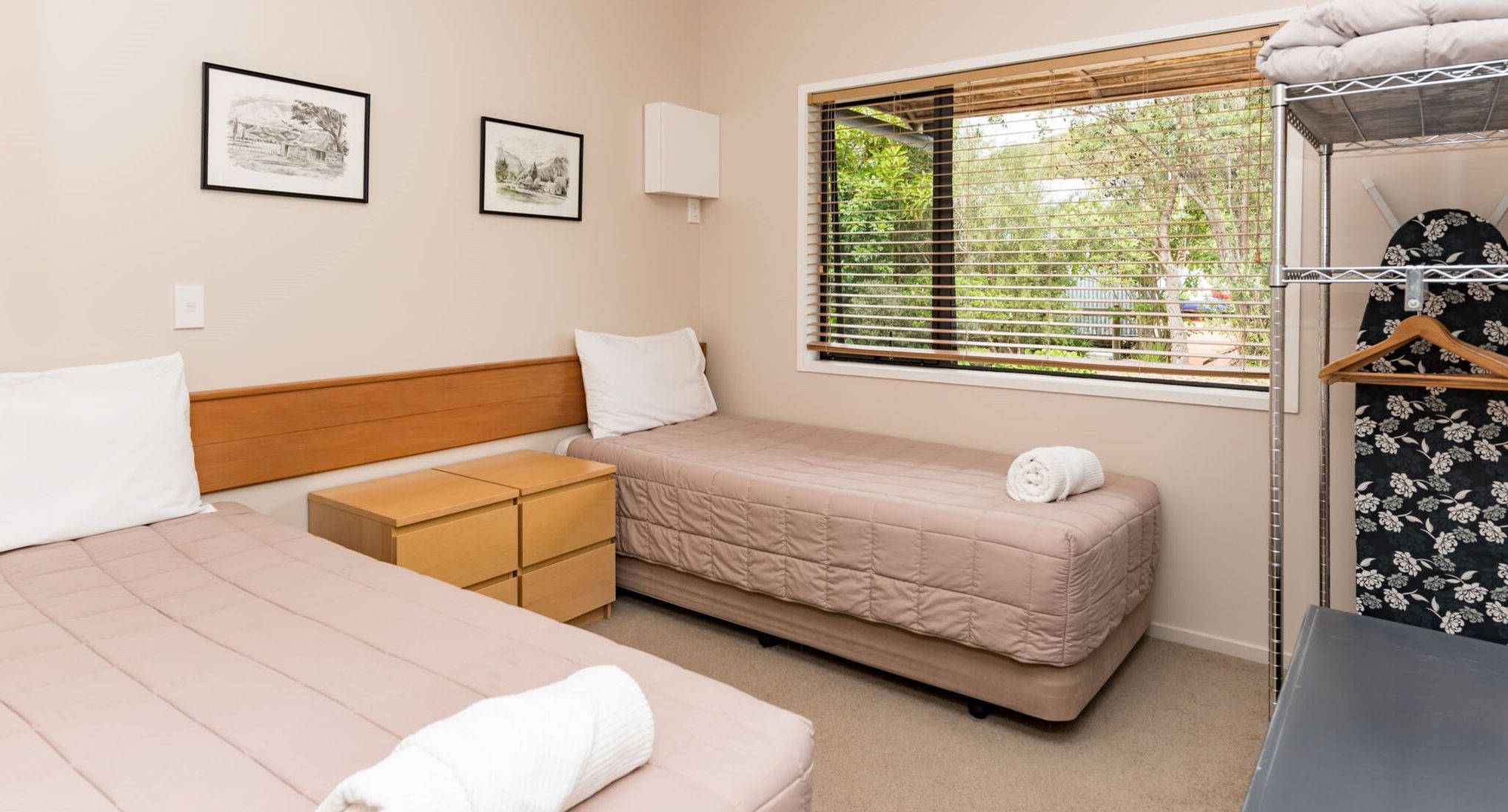 A room with two beds, a wooden headboard, a small wooden cabinet, and a window with blinds.