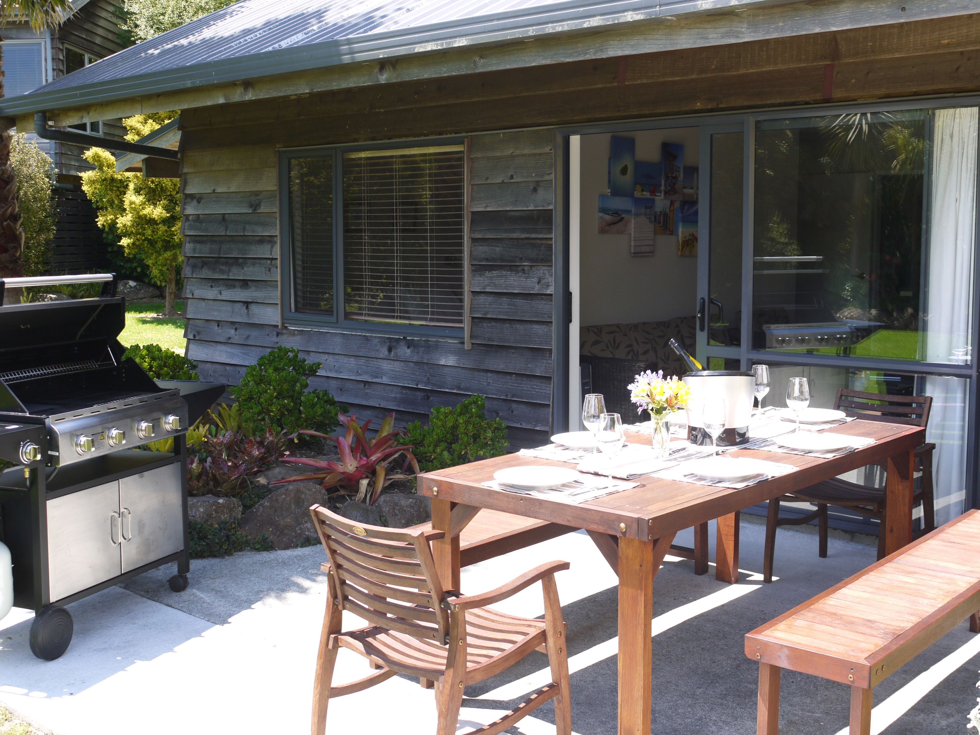 Un espacio de comedor al aire libre con una mesa de madera dispuesta para dos, una parrilla y una ventana con vistas al jardín.