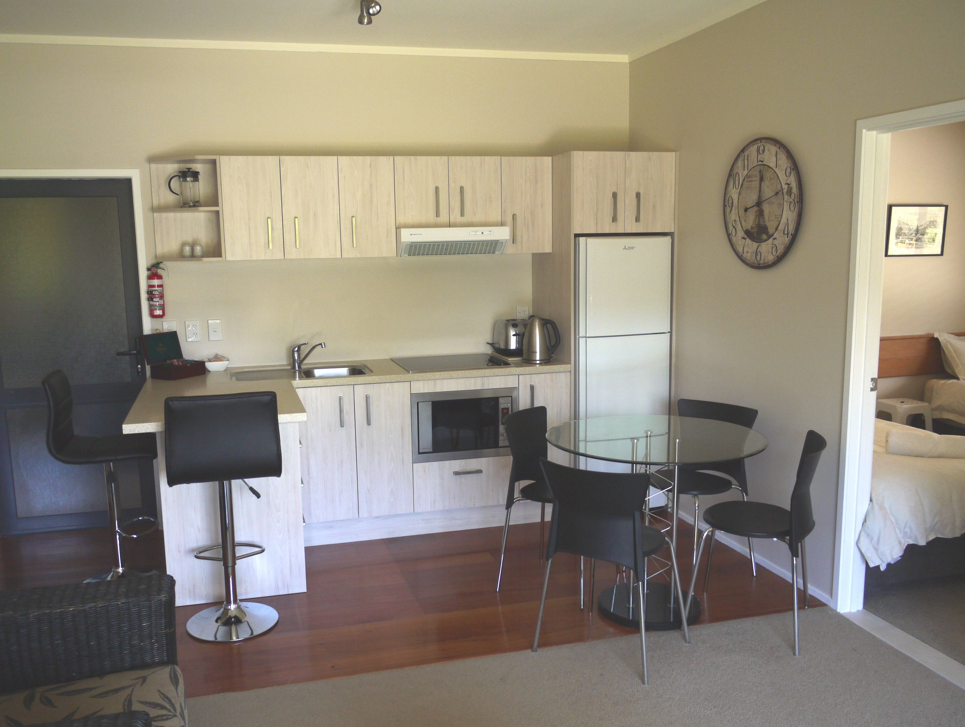 A compact kitchen with a refrigerator, stove, and sink. There are two black bar stools and a glass table. A clock is on the wall.