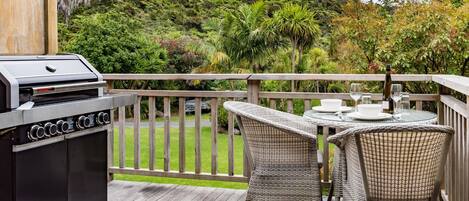 Une terrasse dotée d’un barbecue, de chaises en osier et d’une table dressée pour les repas, offrant une vue sur un jardin luxuriant et une colline boisée.