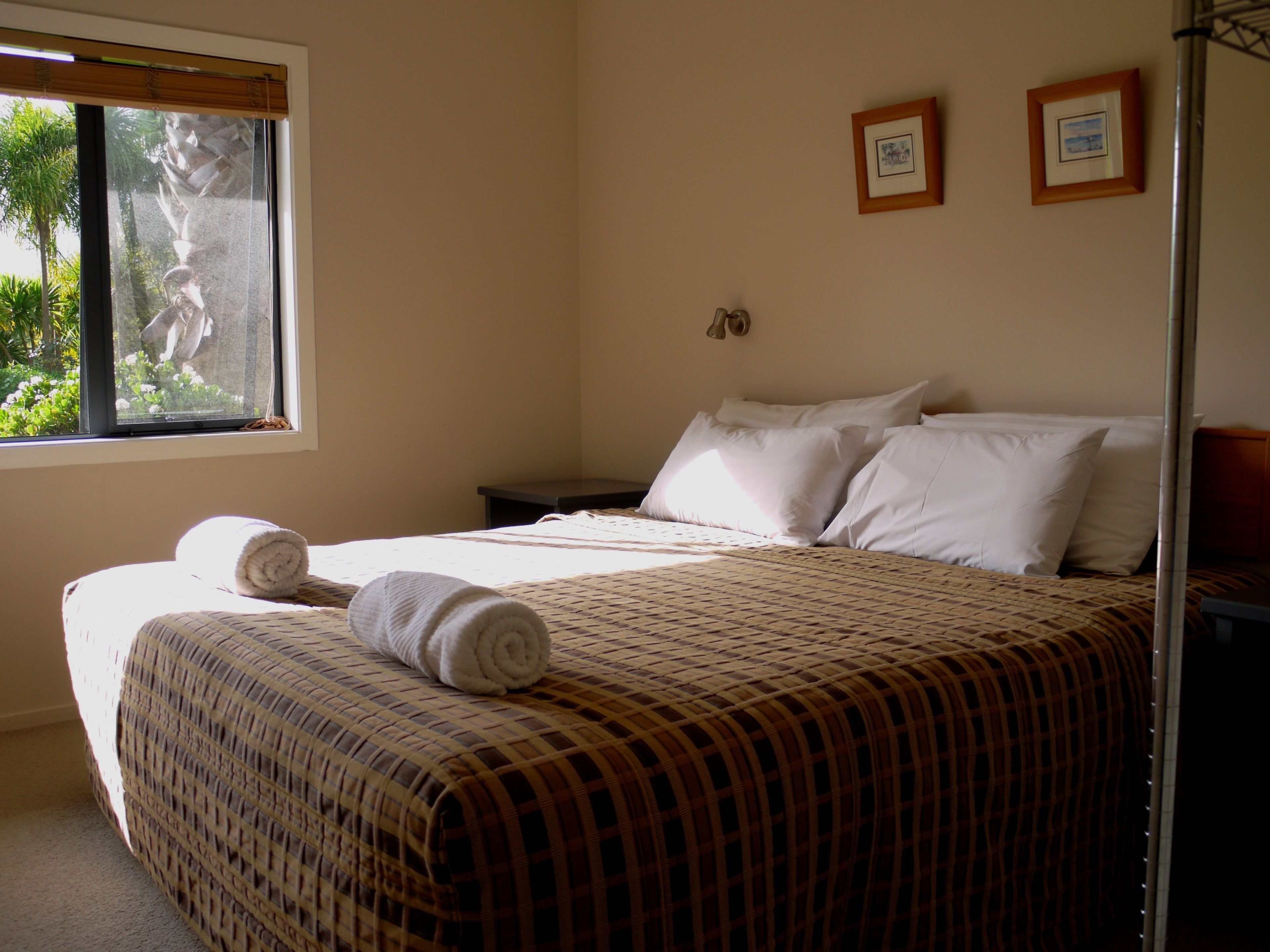 A neatly made bed with a checkered bedspread, two pillows, and two rolled towels. There is a window with a view of greenery outside.