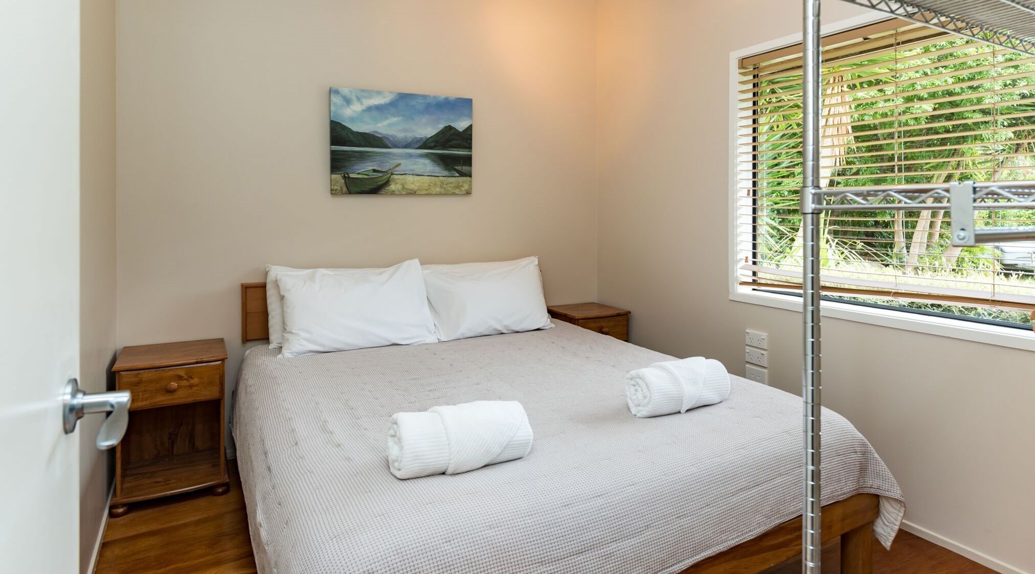 A bedroom with a bed, two pillows, a bedside table, a nightstand, a window with blinds, and a painting on the wall.