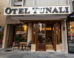 El exterior del OTEL TUNALI, con una entrada destacada con puertas de vidrio, una pequeña zona de estar con dos sillas y una mesa, y un hidrante visible en la acera.