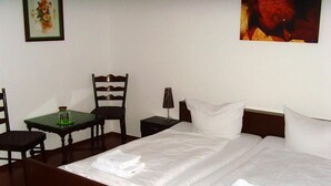 A hotel room with a double bed, a small table with a green tablecloth, two chairs, a nightstand with a lamp, and a framed picture on the wall. - Schlossberg (Heppenheim)
