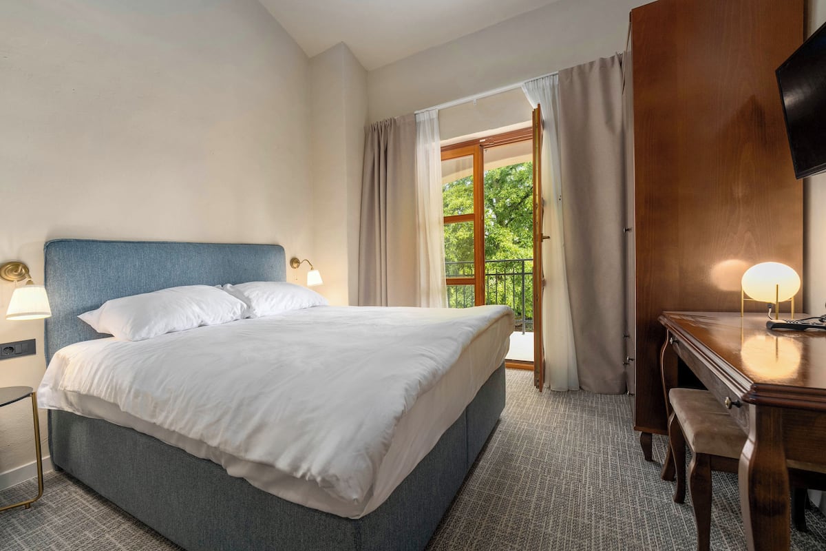 Ein Hotelzimmer mit Bett, Nachttischlampen, einem Schreibtisch mit Stuhl und Blick ins Freie.