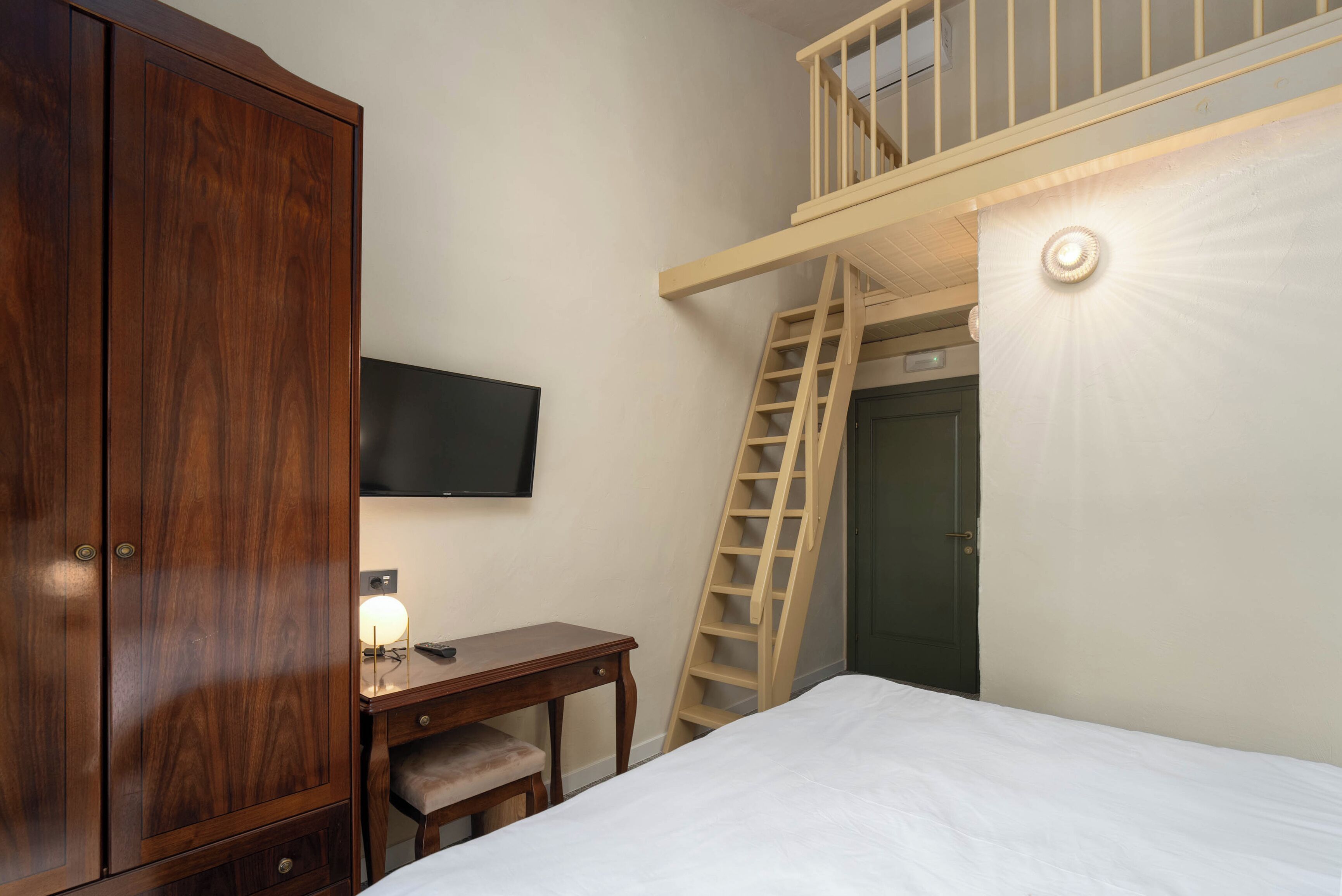 A bedroom with a wooden wardrobe, a desk with a lamp, a bed, and a wooden ladder leading to a loft area.