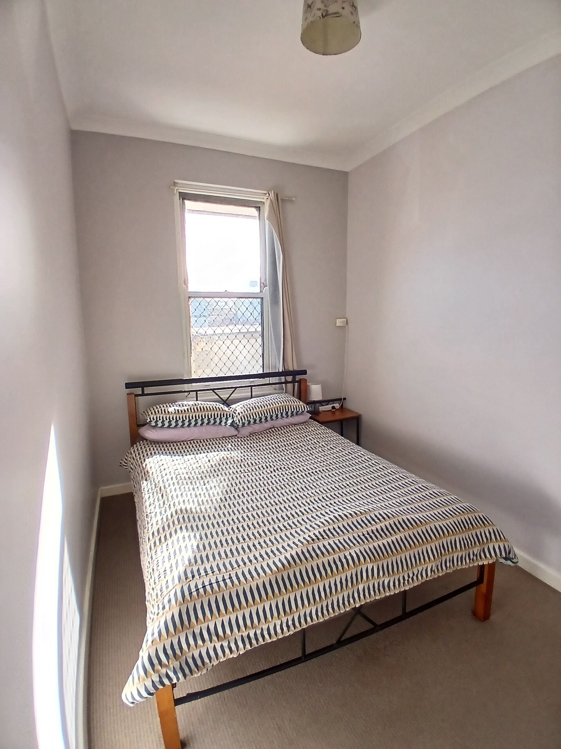 A bedroom with a bed, a window with curtains, a nightstand, and a ceiling light.