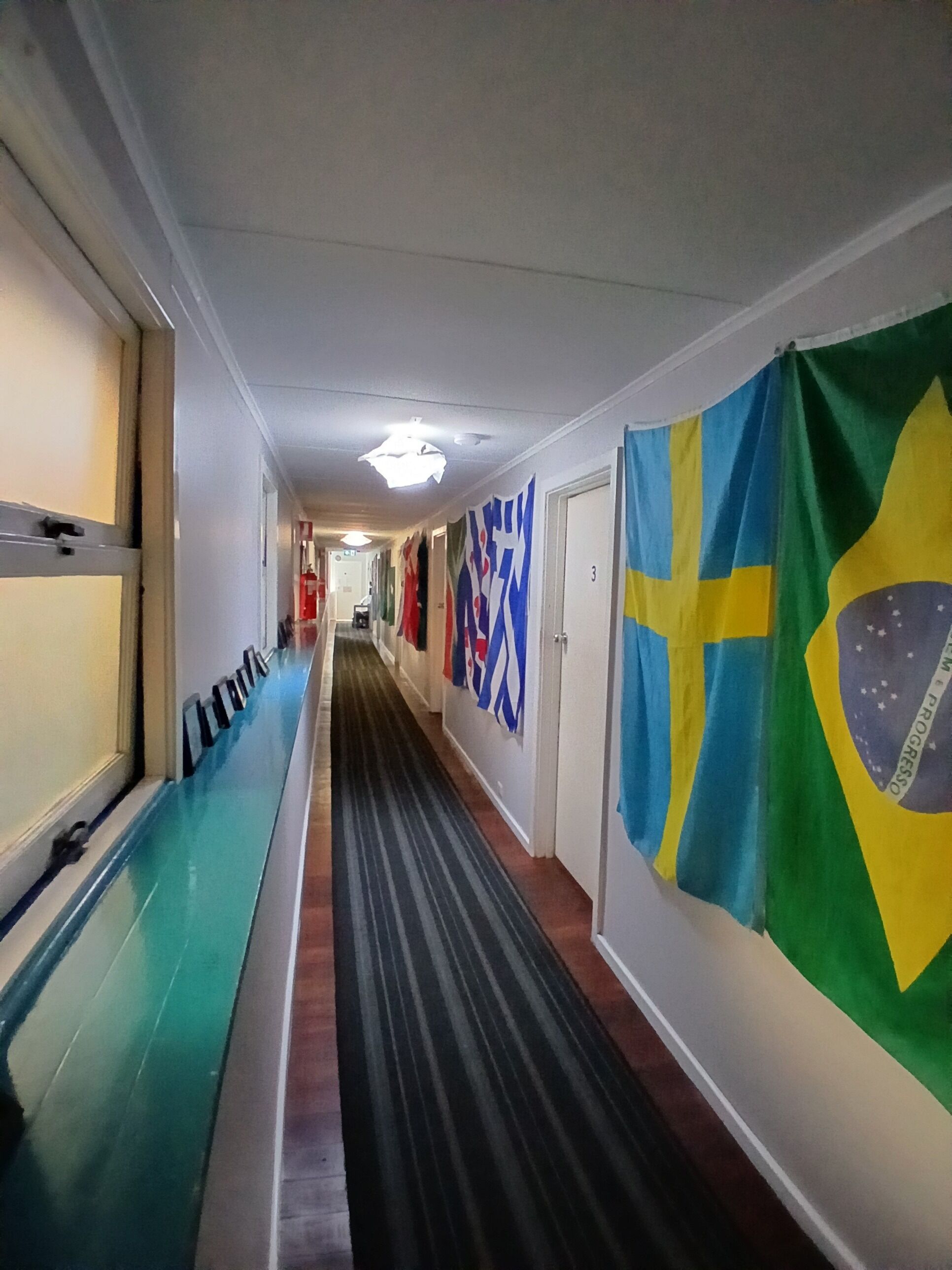 Un couloir avec un tapis à rayures, un drapeau bleu et jaune, et une porte portant le numéro 3.