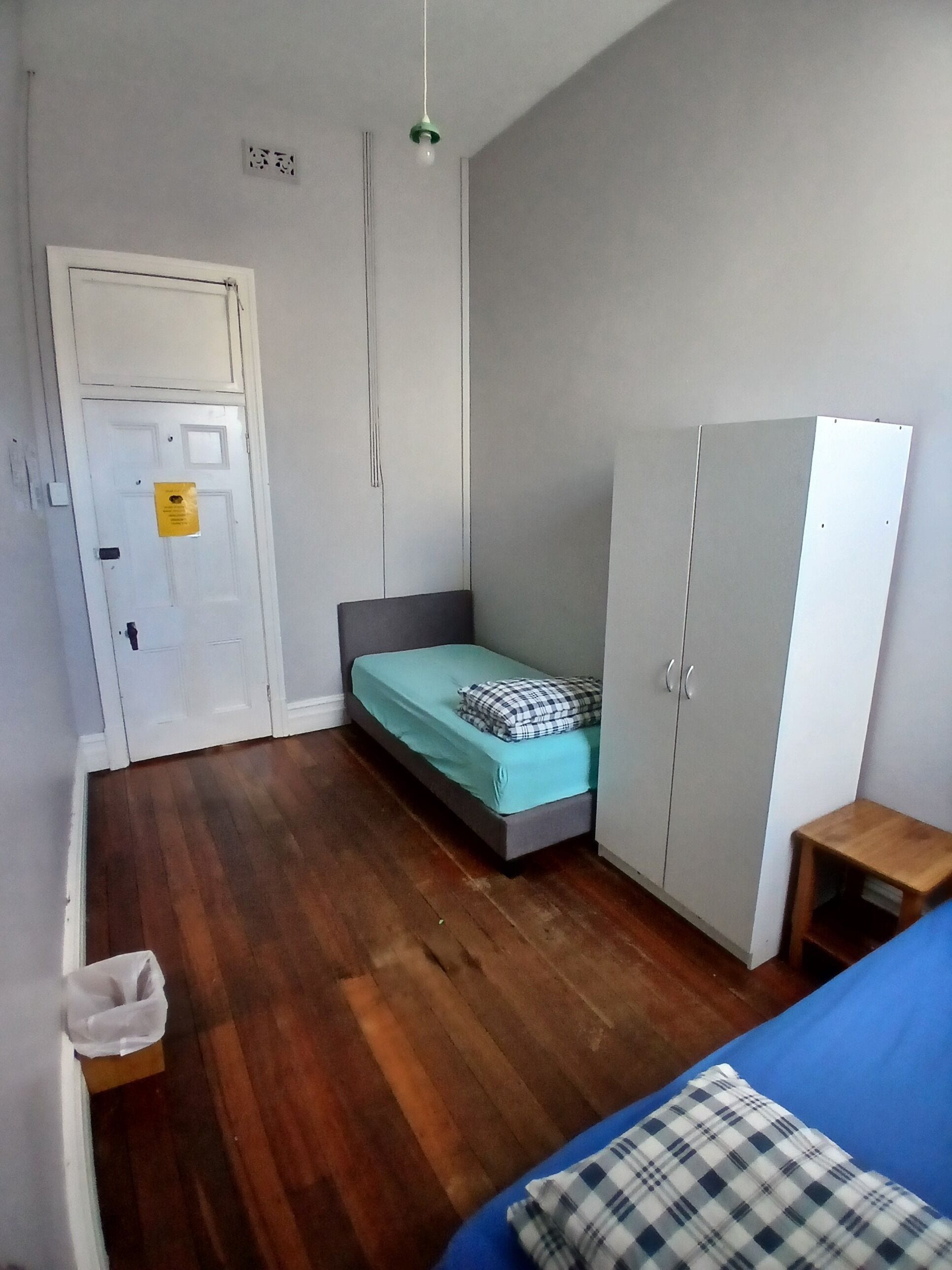 Una habitación pequeña y funcional con una cama individual, un armario, una mesita y una puerta con un cartel.