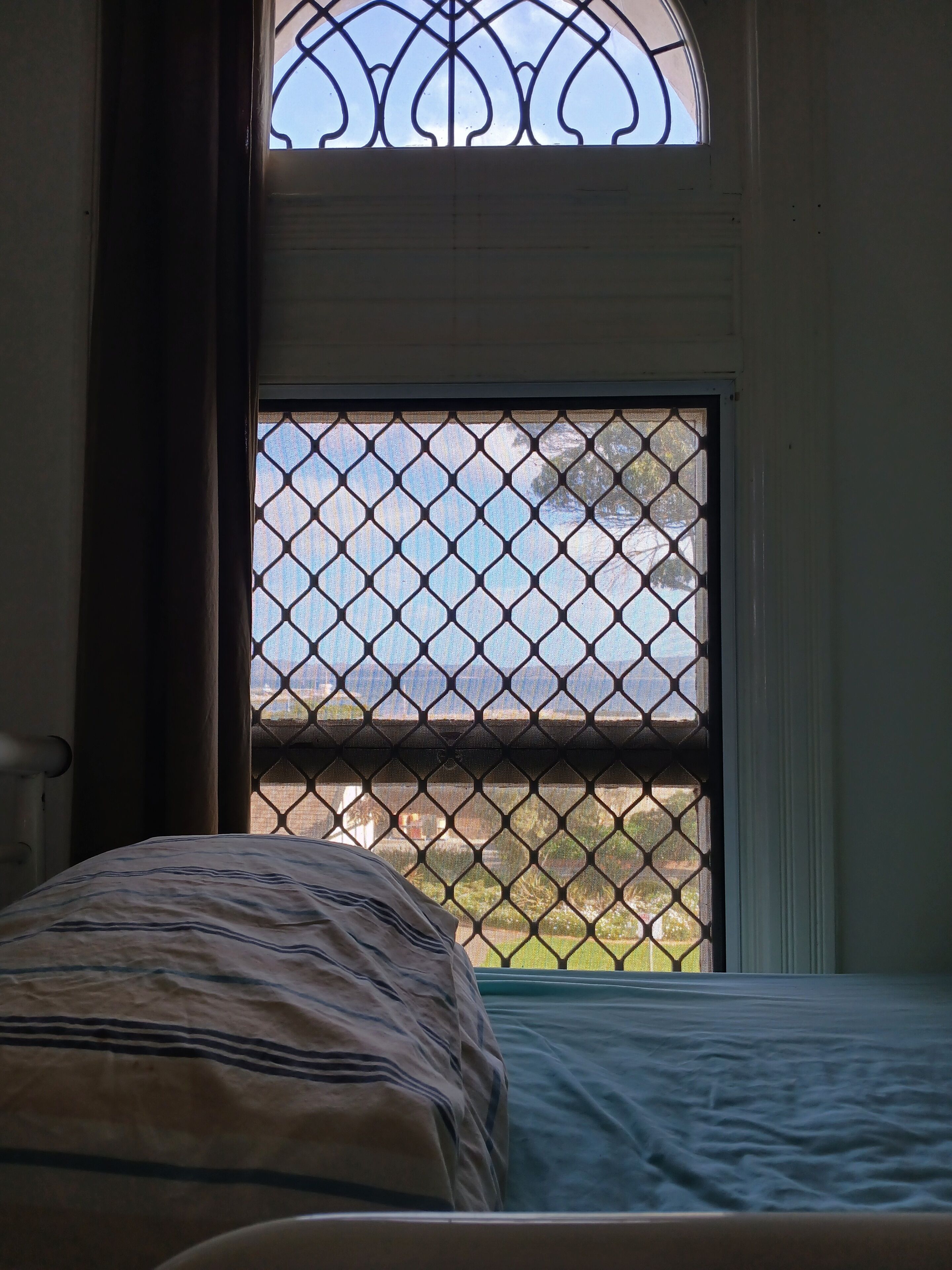 A bedroom with a bed, a window with a metal screen, and a view of the outdoors.