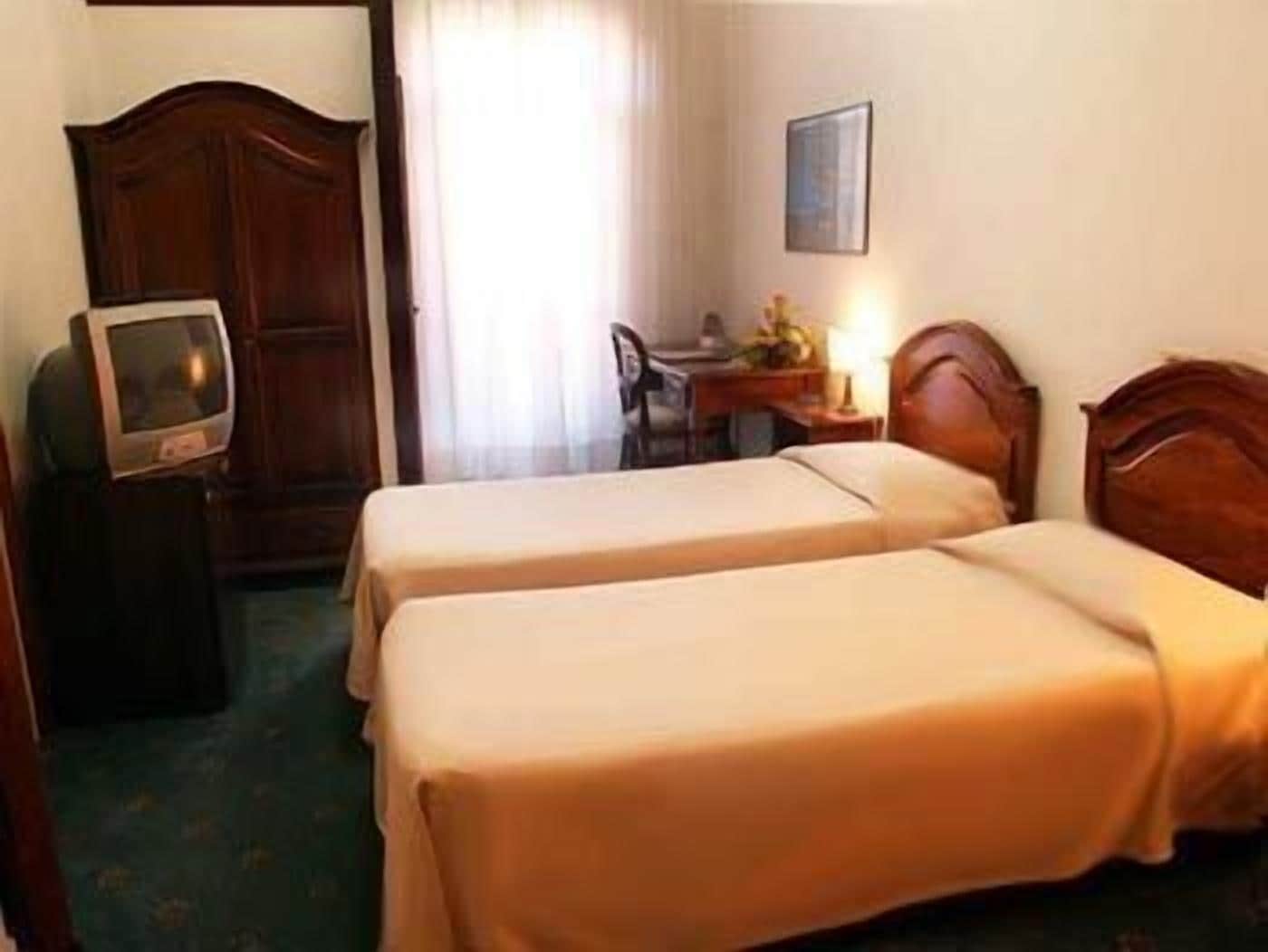 A hotel room with two single beds, a wooden wardrobe, a desk with a chair, a television, and a framed picture on the wall.