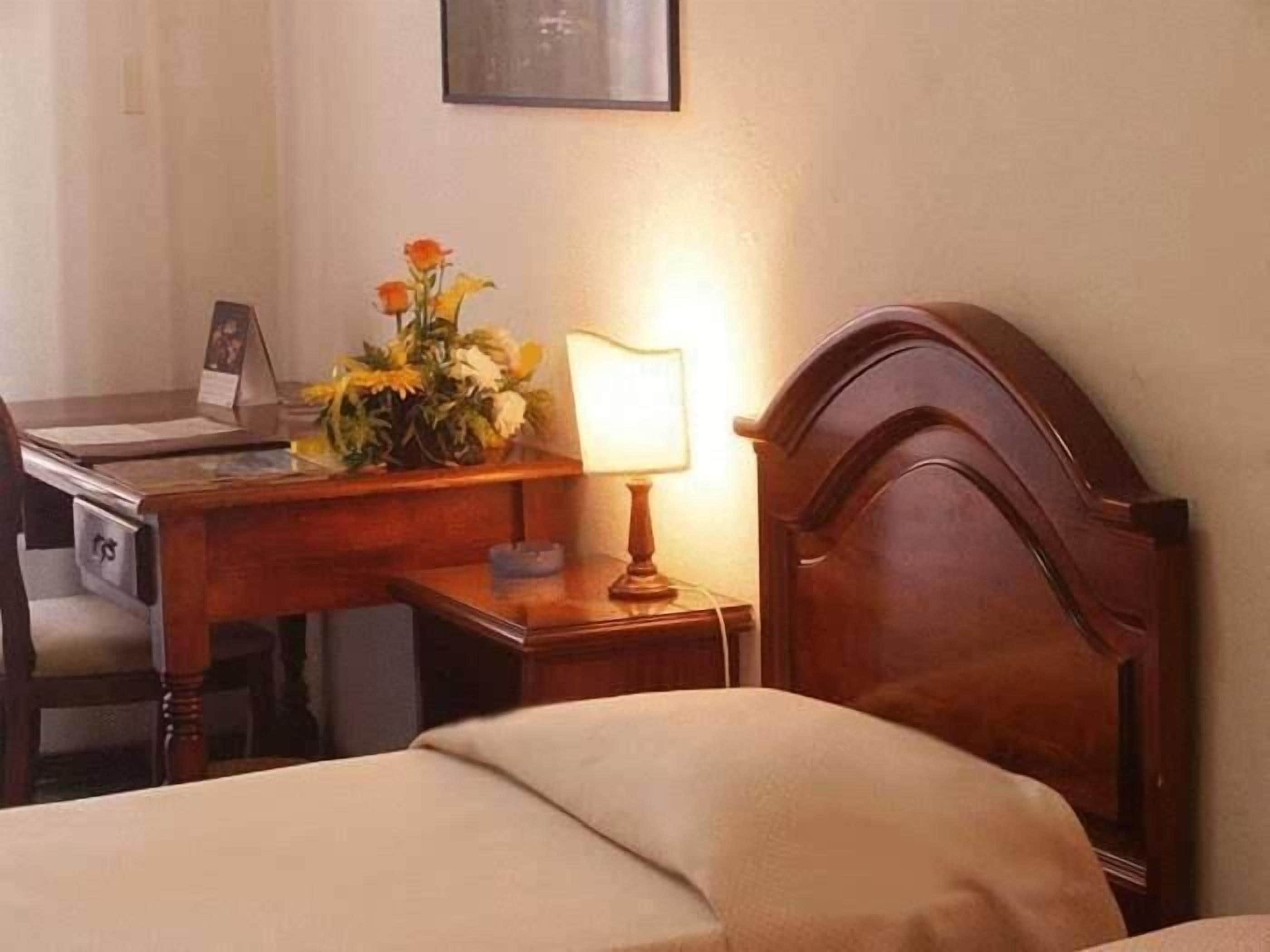 Habitación de hotel con un escritorio de madera, una lámpara, una cama y una mesita de noche con un jarrón de flores.