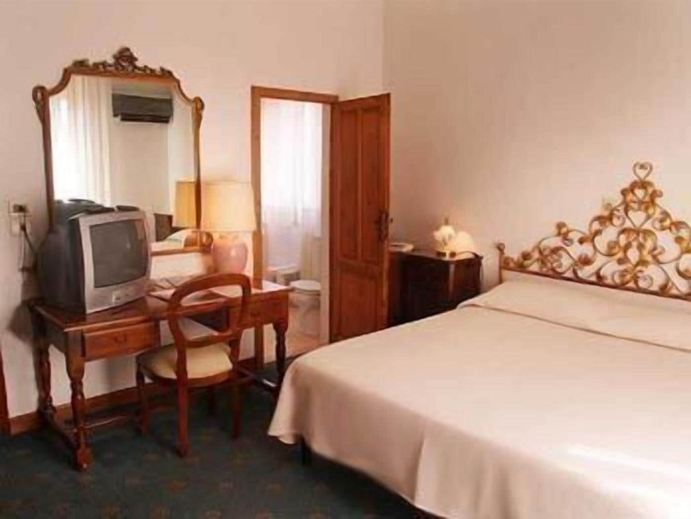 Habitación de hotel con cama, escritorio con televisor, una silla, un espejo y mesita de noche con lámpara.