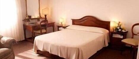 A hotel room with a single bed, a desk, a chair, and a nightstand with a lamp.