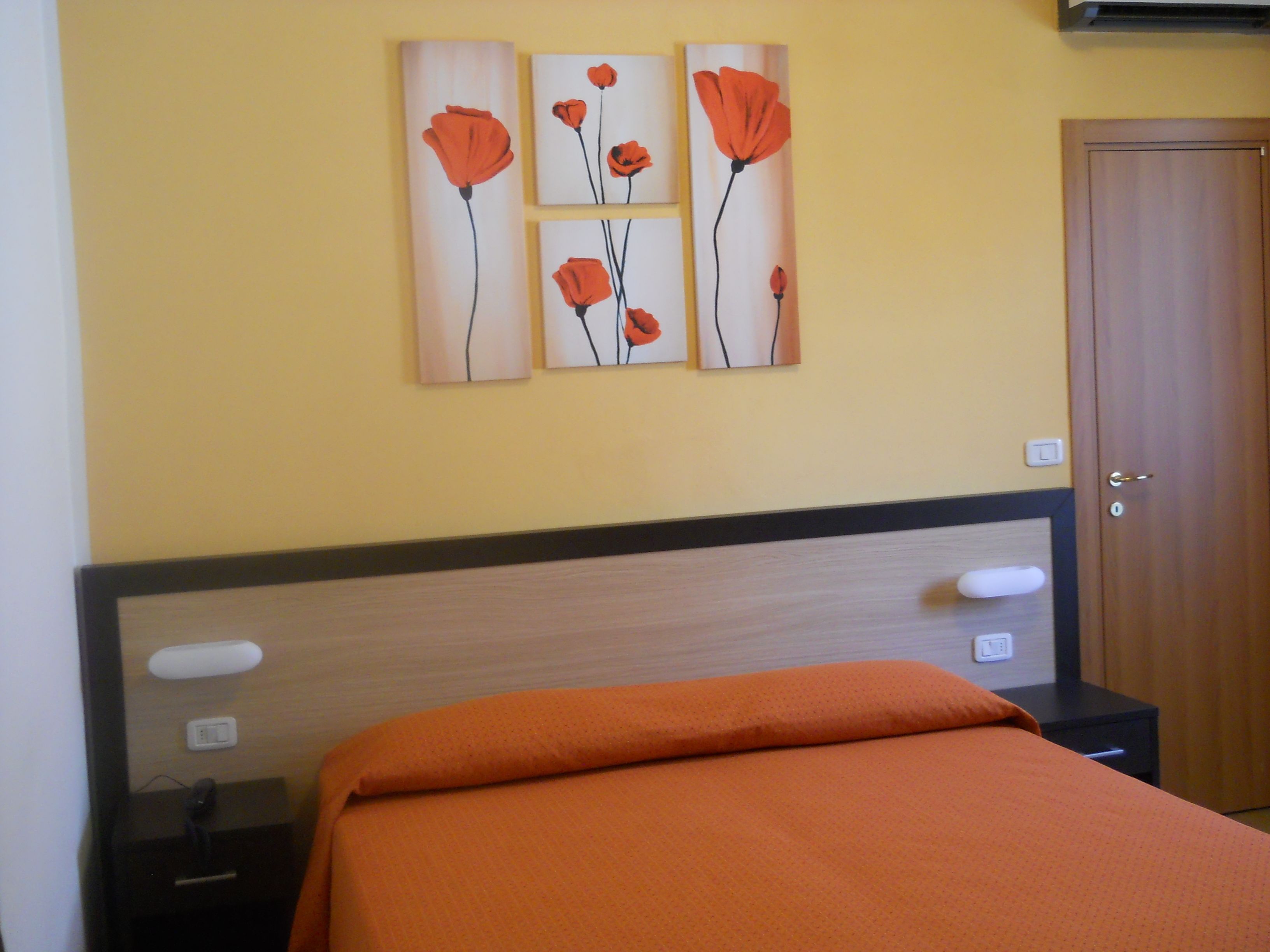 A bedroom with a bed, a nightstand, a wall-mounted artwork of red flowers, and a door.