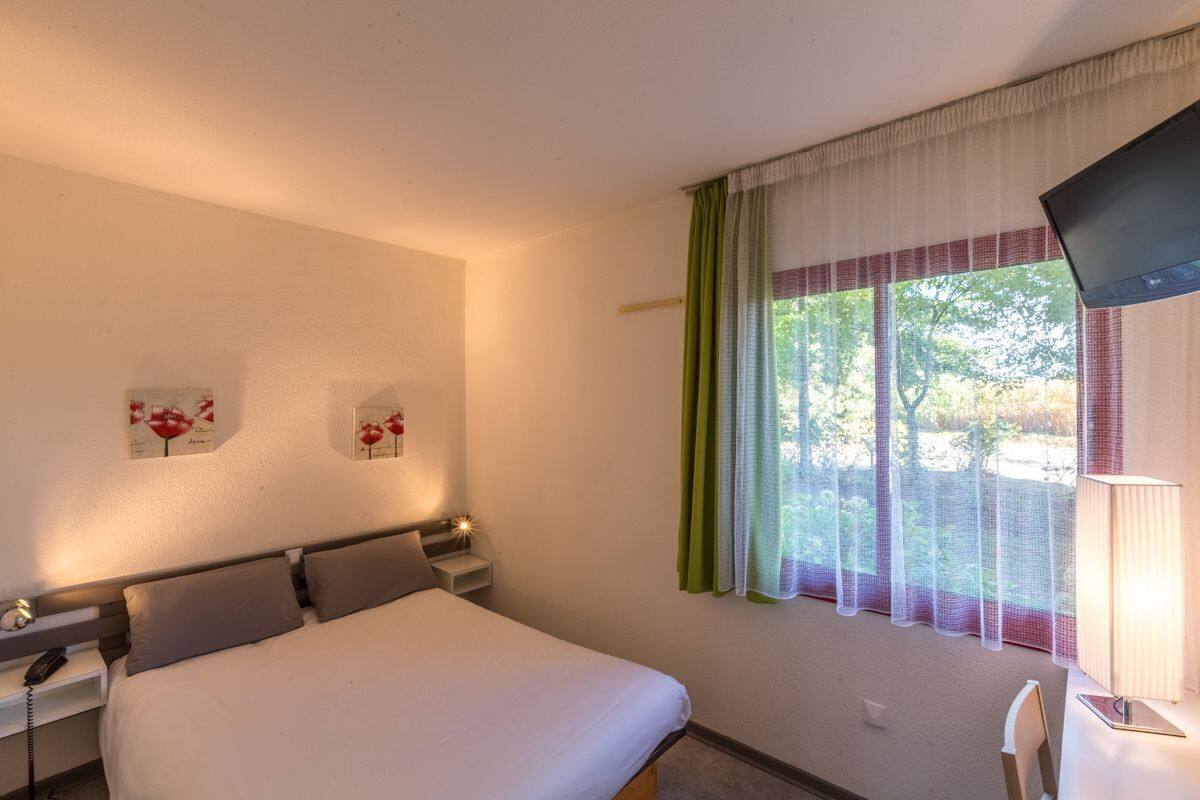 Une chambre d’hôtel avec un lit, une table de chevet, une lampe, une télévision et une fenêtre avec des rideaux.