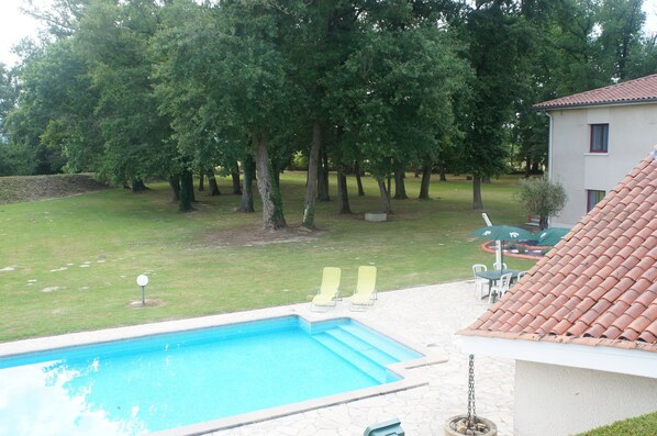 A swimming pool with two sun loungers, surrounded by trees and a building with a tiled roof.