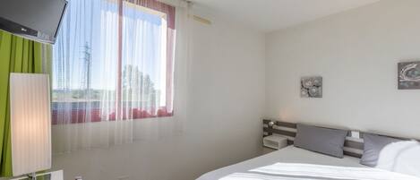Un dormitorio con cama, televisor montado en la pared, ventana con cortinas y mesita de noche con lámpara.