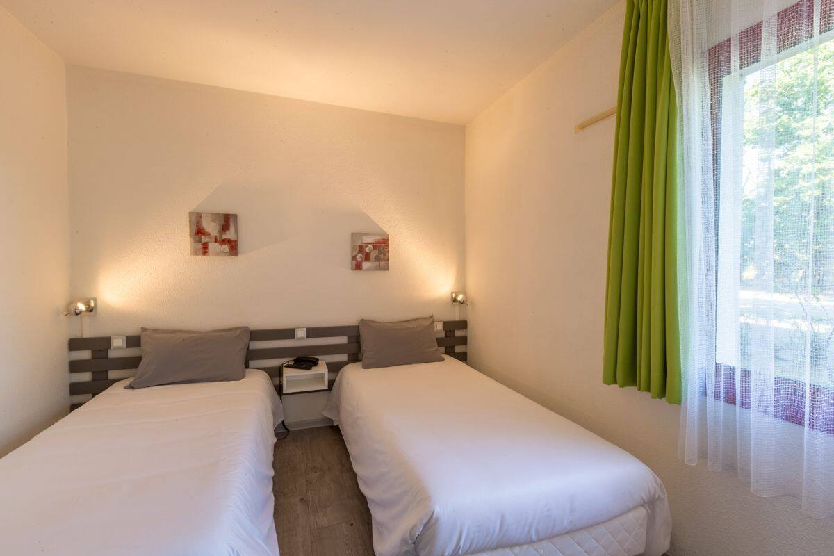 Une chambre double avec deux lits, une table de chevet, un téléphone fixé au mur et une fenêtre avec des rideaux verts.