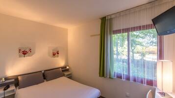 A hotel room with a bed, bedside table, lamp, TV, and a window with curtains.