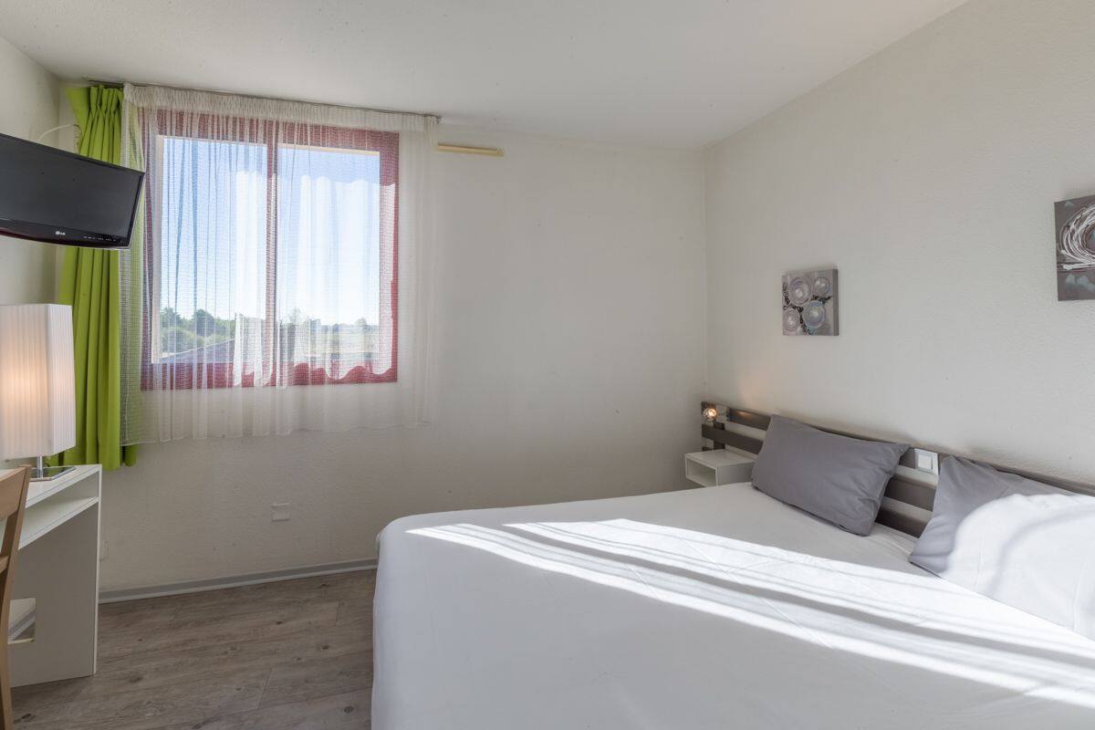 Une chambre à coucher avec un lit, un bureau, une télévision et une fenêtre avec des rideaux.