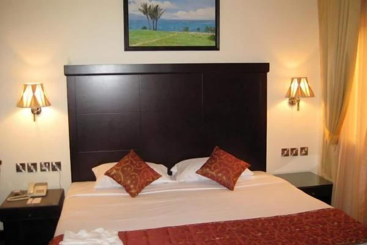 A hotel room with a large bed, two wall-mounted lamps, a painting of a landscape, and a telephone on a side table.
