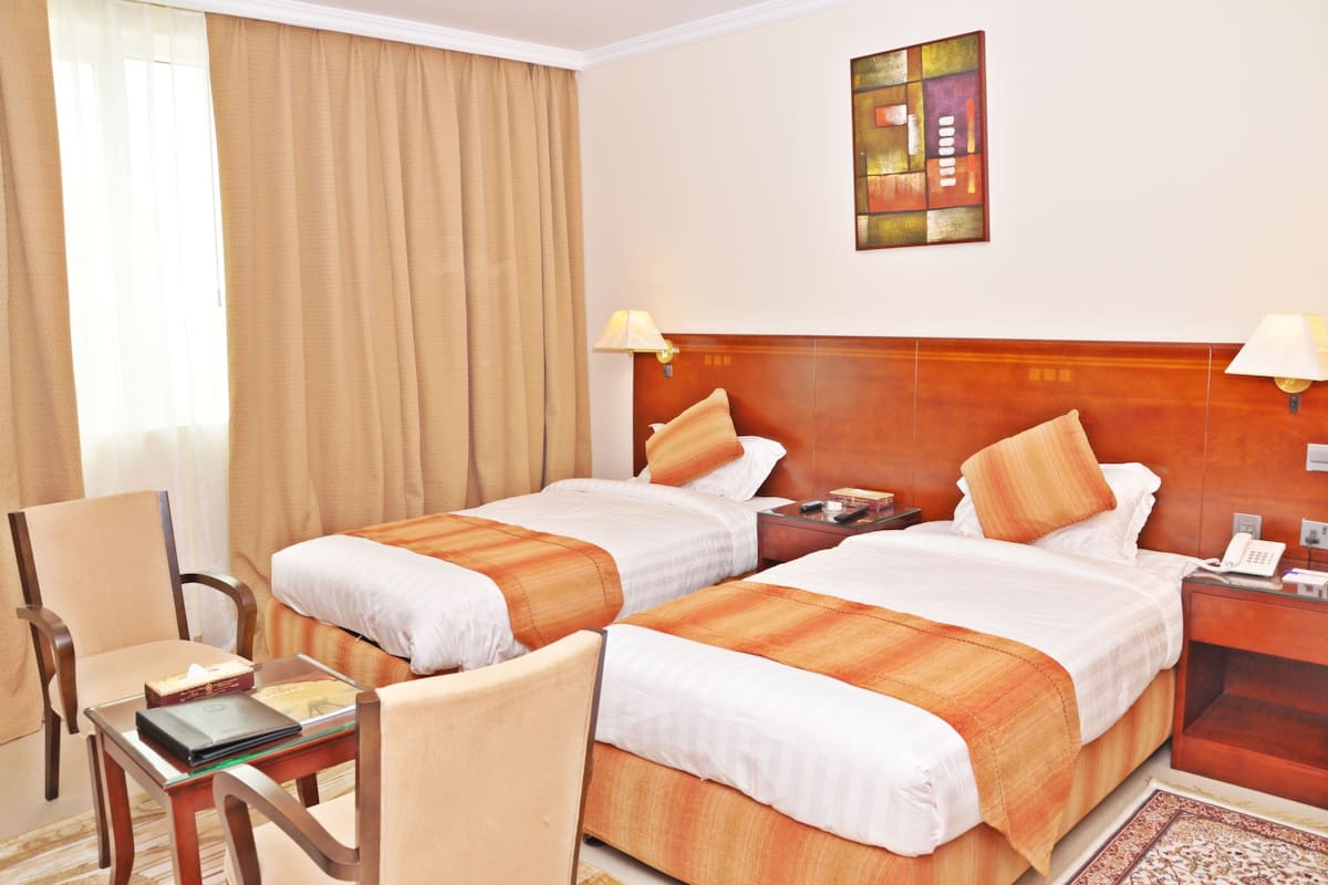 A hotel room with two beds, a desk, a chair, and a nightstand with a lamp.